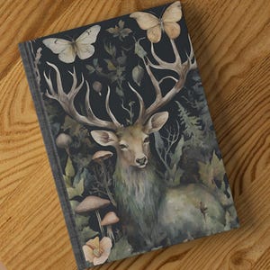 Può includere: Un diario con copertina rigida con un'illustrazione ad acquerello di un cervo con grandi corna, farfalle, funghi e fogliame. La copertina ha uno sfondo scuro con un tema forestale, su una superficie di legno.