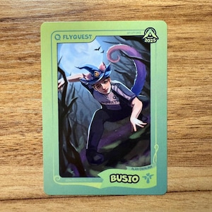 A trading card featuring lolesports LCS and LEC professional player Alan Cwalina, aka Busio, in the FlyQuest 2025 pink jersey. He is drawn landing on the ground of a dark spooky forest as if he just ulted as Bewitching Neeko, one of his iconic League of Legends champions. The card has a green gradient border with the text "BUSIO" and the Support icon at the bottom right, 'BEWITCHING NEEKO' at the bottom left, the LTA North logo and 2025 at top right, and the FLYQUEST name and logo at top left.