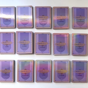 May include: A set of 15 holographic trading cards featuring various characters from the Disney movie "Descendants". Each card has a purple background with a gold border and features a character from the movie. The cards are arranged in three rows of five.