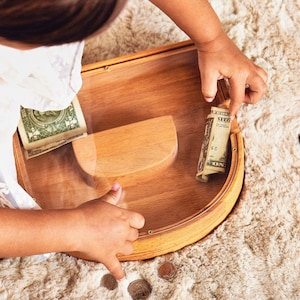 K&ouml;nnte beinhalten: Eine Holzspardose mit einer transparenten Acrylfront, die gerollte und gefaltete Dollarnoten enth&auml;lt. Ein Kind interagiert mit der Box, M&uuml;nzen sind in der N&auml;he verstreut. Die Box hat ein geschwungenes Design und eine helle Holzoberfl&auml;che.