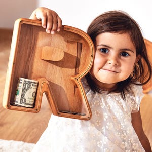 May include: A wooden money box shaped like the letter R. The money box is made of natural wood and has a slot for coins on the side. A child is holding the money box in front of their face. The money box has a few coins and a dollar bill inside.