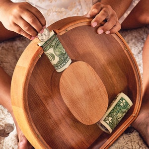 May include: A wooden coin bank with a clear lid, showing rolled up dollar bills inside. The bank is shaped like a bowl and has a natural wood finish.