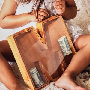 May include: A wooden money box shaped like the letter M with a clear plastic front. The money box is filled with rolled up pound notes.