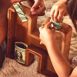 May include: A wooden money box with slots for bills. A child is putting a dollar bill into the box.