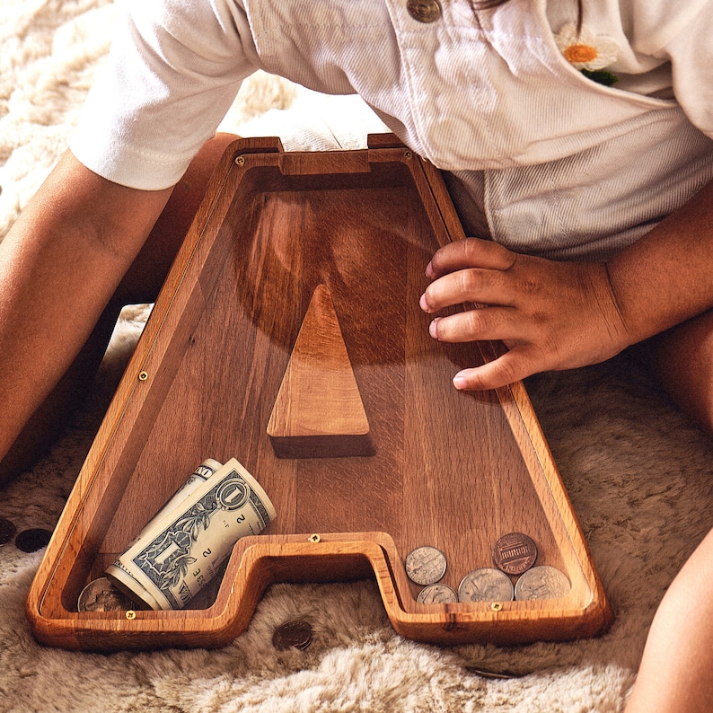 K&ouml;nnte beinhalten: Holzsparb&uuml;chse in Form des Buchstabens "A" mit klarer Vorderseite. Die Bank enth&auml;lt einen aufgerollten Dollarschein und mehrere M&uuml;nzen. Die Bank ist aus hellbraunem Holz gefertigt und hat einen dreieckigen Holzakzent.