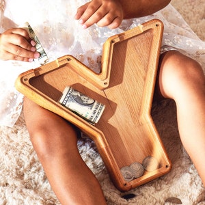 May include: A wooden Y-shaped money box with a slot for coins and notes. A child is putting money into the box.