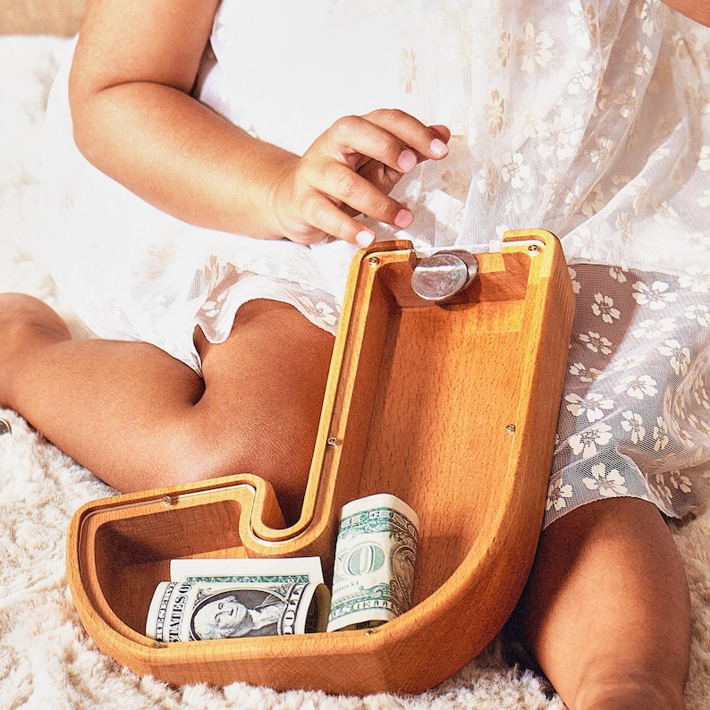 May include: A wooden J-shaped money box with a coin slot and a compartment for storing money. The money box is made of light brown wood and has a natural finish. There are two dollar bills inside the money box.