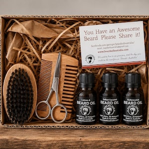 May include: A cardboard gift box containing a wooden beard brush with black bristles, a wooden comb, silver metal grooming scissors, and three small amber glass bottles of beard oil labelled Lemon, Salted Caramel, and Butterscotch.