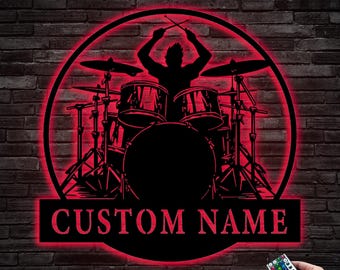 May include: A black metal silhouette of a drummer playing a drum set with a red LED light behind it. The silhouette is in a circle with the words "CUSTOM NAME" below it.