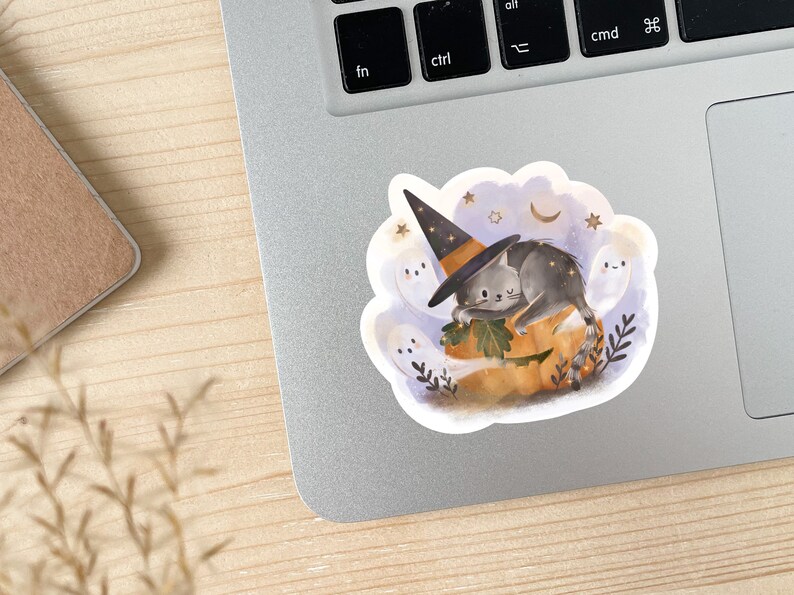 May include: A Halloween-themed sticker featuring a gray cat wearing a witch hat, resting on a carved pumpkin. The sticker includes ghosts, stars, and a crescent moon, all set against a light blue background. The design has a watercolor style.