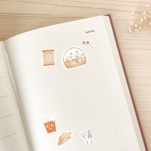 May include: Open notebook with stickers of gardening tools and flowers. Stickers include a seed packet, a basket of flowers, a watering can, a sun hat, and gardening tools. The notebook is open to a dotted page with the word "NOTES" at the top.