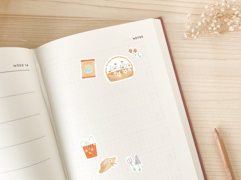 May include: Open notebook with stickers of gardening tools and flowers. Stickers include a seed packet, a basket of flowers, a watering can, a sun hat, and gardening tools. The notebook is open to a dotted page with the word "NOTES" at the top.