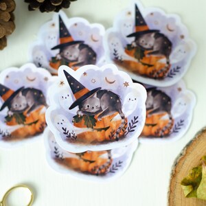 May include: A collection of Halloween-themed stickers featuring a gray cat wearing a witch hat, resting on a carved pumpkin. The stickers have a cloud-like border with stars and ghosts. The color palette includes orange, gray, and purple.