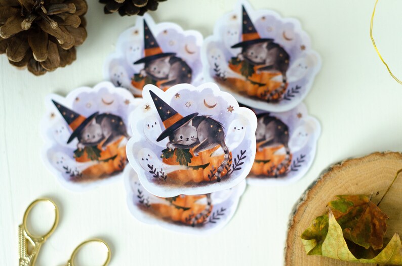 May include: A collection of Halloween-themed stickers featuring a gray cat wearing a witch hat, resting on a carved pumpkin. The stickers have a cloud-like border with stars and ghosts. The color palette includes orange, gray, and purple.