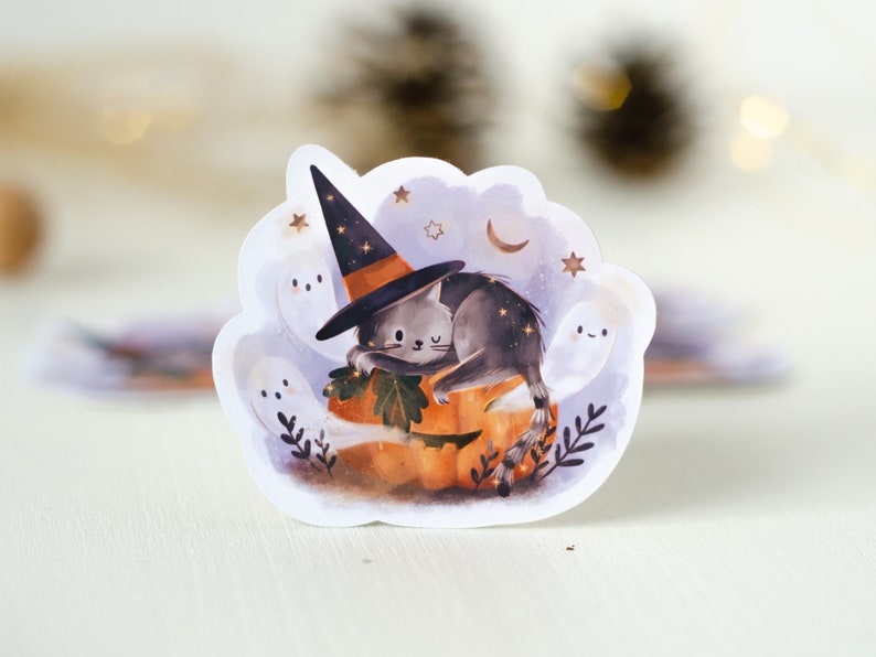 May include: A whimsical Halloween sticker featuring a gray cat wearing a witch hat, resting on an orange pumpkin. The design includes friendly ghosts, stars, and a crescent moon, creating a spooky yet cute aesthetic.