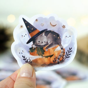 May include: A Halloween-themed sticker featuring a gray cat wearing a witch hat, resting on a carved pumpkin. The sticker includes ghosts, stars, and a crescent moon, with a whimsical, illustrated style.