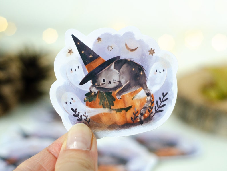 May include: A Halloween-themed sticker featuring a gray cat wearing a witch hat, resting on a carved pumpkin. The sticker includes ghosts, stars, and a crescent moon, with a whimsical, illustrated style.