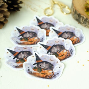 May include: A collection of Halloween-themed stickers featuring a gray cat wearing a witch hat, resting on a pumpkin. The stickers have a whimsical design with ghosts, moons, and stars, perfect for seasonal crafts.