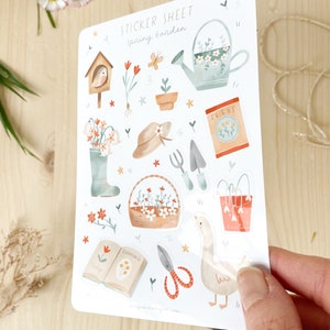 May include: A sticker sheet with a spring garden theme. The sheet features illustrations of gardening tools, flowers, a watering can, a birdhouse, and a goose. The text "STICKER SHEET Spring Garden" is at the top. The stickers are in soft pastel colours.