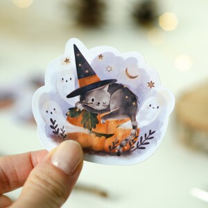 May include: A Halloween-themed sticker featuring a gray cat wearing a witch hat, resting on an orange pumpkin. The sticker includes white ghosts, stars, and a crescent moon, creating a whimsical, spooky design. The sticker is held by a hand.