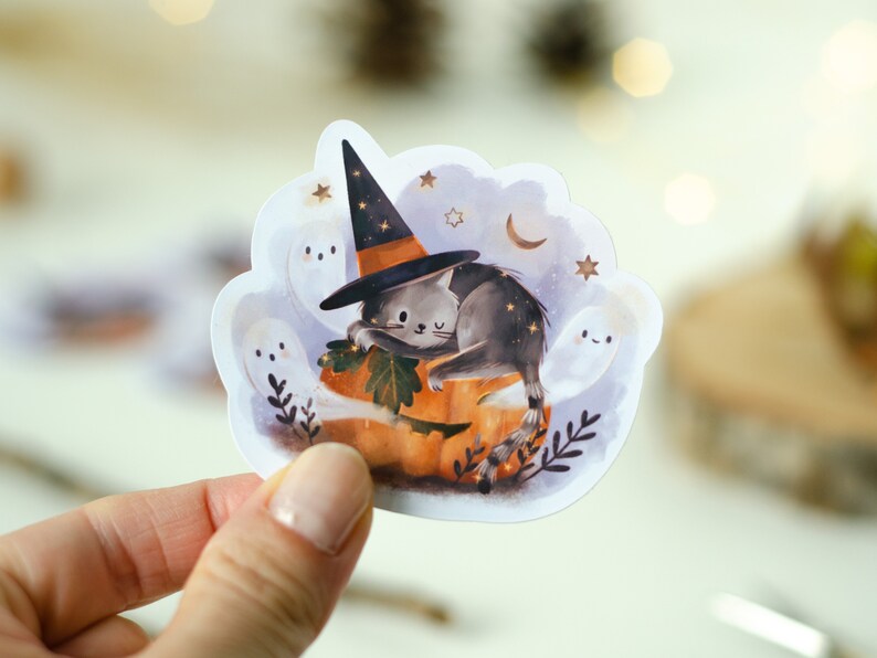 May include: A Halloween-themed sticker featuring a gray cat wearing a witch hat, resting on an orange pumpkin. The sticker includes white ghosts, stars, and a crescent moon, creating a whimsical, spooky design. The sticker is held by a hand.