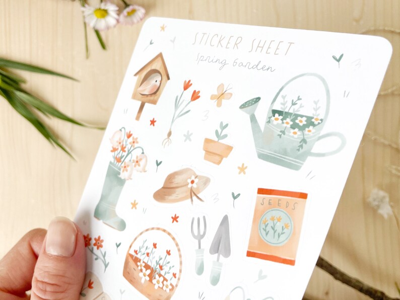 May include: A sticker sheet featuring watercolour illustrations of garden-themed items, including a birdhouse, flowers, a watering can, and gardening tools. The text "STICKER SHEET Spring Garden" is at the top.