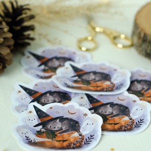 May include: A collection of stickers featuring a gray cat wearing a witch hat, sleeping on an orange pumpkin. The stickers have a white cloud-shaped border with stars, moons, and smiling ghosts. The overall theme is Halloween.