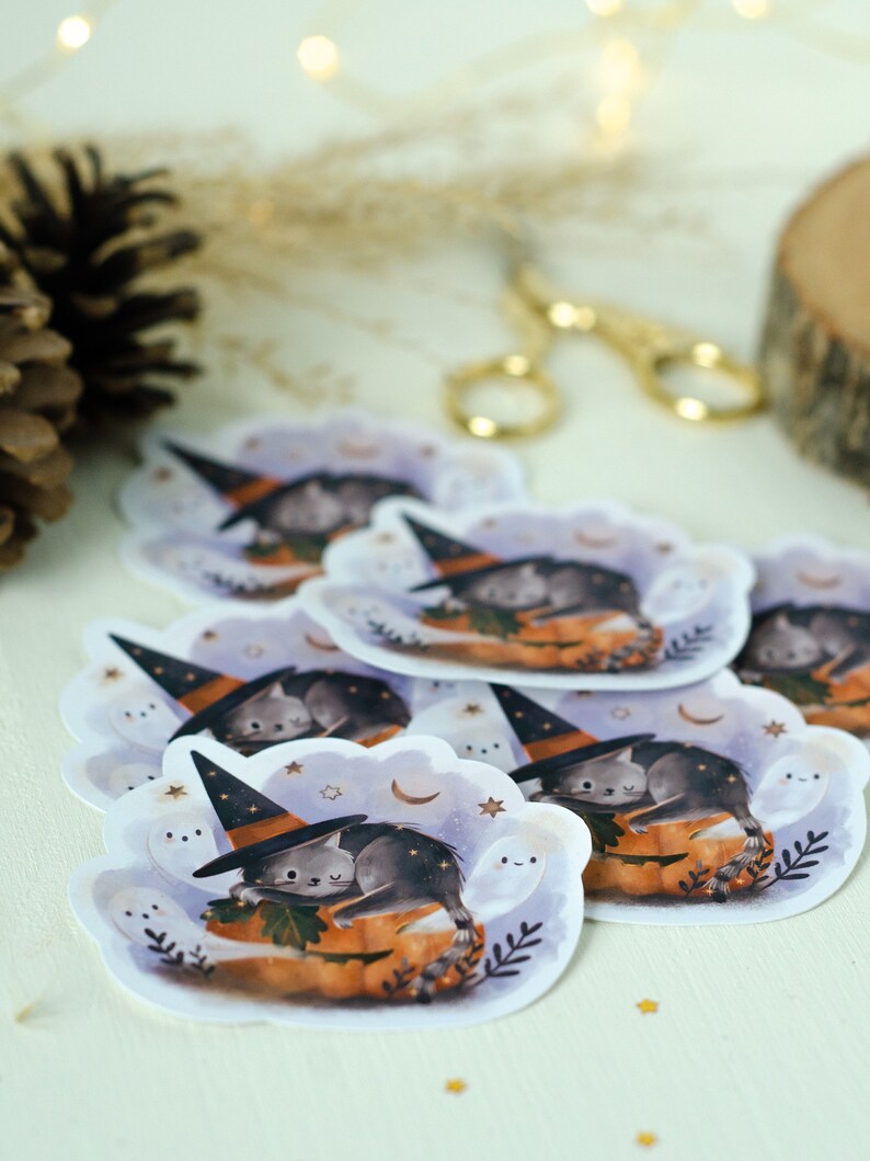 May include: A collection of stickers featuring a gray cat wearing a witch hat, sleeping on an orange pumpkin. The stickers have a white cloud-shaped border with stars, moons, and smiling ghosts. The overall theme is Halloween.