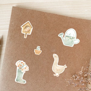 May include: A selection of watercolour stickers with garden-themed illustrations. Includes a birdhouse with a bird, a watering can with flowers, a potted plant, a goose, and a wellington boot filled with flowers. The stickers are on a brown surface.