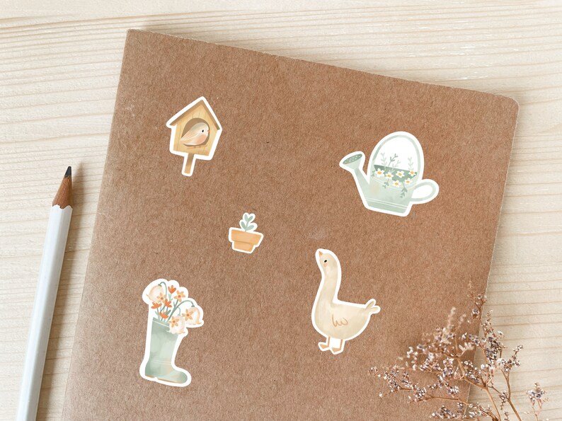 May include: A selection of watercolour stickers with garden-themed illustrations. Includes a birdhouse with a bird, a watering can with flowers, a potted plant, a goose, and a wellington boot filled with flowers. The stickers are on a brown surface.