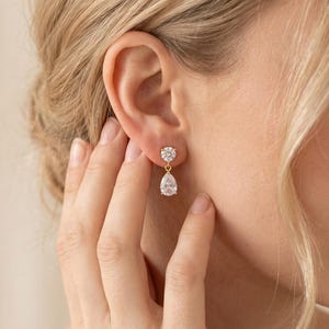 May include: A gold drop earring featuring a small round brilliant-cut clear stone at the top, connected by a thin gold link to a larger pear-shaped clear stone pendant. The earring is crafted with a polished gold setting and secure post back.