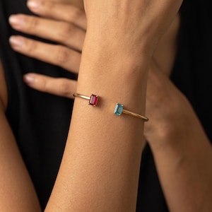 May include: A gold-toned open cuff bracelet featuring two rectangular-cut gemstones. One end of the bracelet is set with a vibrant red gemstone, while the opposite end is set with a clear, light blue gemstone, creating a contrasting color design.