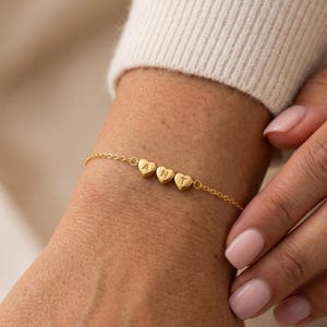 Puede incluir: Una pulsera dorada con dijes en forma de corazón que deletrean "ANT". La pulsera se lleva en la muñeca, con un suéter color crema en el fondo. La joya es delicada y elegante.