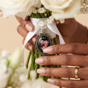 Peut inclure: Un médaillon photo ovale en métal couleur bronze avec le portrait de deux personnes, fixé sur un ruban de satin blanc entourant un bouquet de roses blanches et de gypsophile. Le cadre vintage présente un motif de bordure en filigrane.