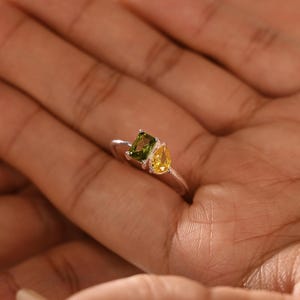 Puede incluir: Un anillo de plata con una gema verde cuadrada y una gema amarilla en forma de pera. El anillo se sostiene en una mano abierta, mostrando los colores vibrantes de las gemas y el delicado diseño del anillo. Es una joya.