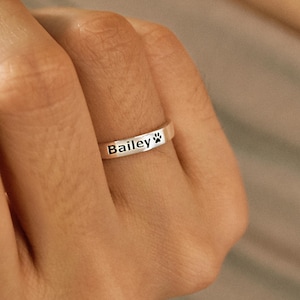 Puede incluir: Un anillo de plata con el nombre "Bailey" grabado y una huella de pata. El anillo tiene un diseño de banda simple, adecuado para el uso diario. El anillo se muestra en un dedo.
