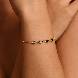 Könnte beinhalten: Ein zartes goldenes Armband mit einer Reihe farbiger, tropfenförmiger Edelsteine. Das Armband enthält klare, grüne, rote und smaragdgrüne Steine. Das Armband wird am Handgelenk getragen.
