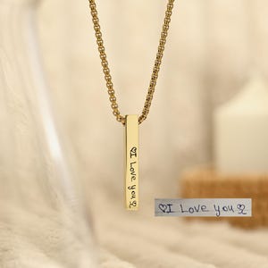 Peut inclure: Un collier pendentif à barre verticale de couleur or avec une chaîne de type boîte. Le pendentif porte l'inscription manuscrite "I Love you". Une petite note manuscrite avec le même texte est à côté du collier.