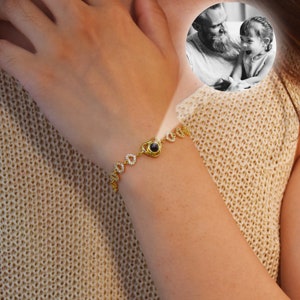 May include: A gold bracelet with a heart-shaped charm that reveals a hidden image when viewed through a small lens. The charm is adorned with small clear stones. The bracelet is on a person's wrist.