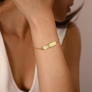 K&ouml;nnte beinhalten: Ein goldfarbenes Armband mit einem gravierten rechteckigen Anh&auml;nger mit hieroglyphischen Symbolen. Das Armband ist an einer zarten Kette befestigt und wird am Handgelenk getragen. Der Schmuck wird vor einem neutralen Hintergrund pr&auml;sentiert.