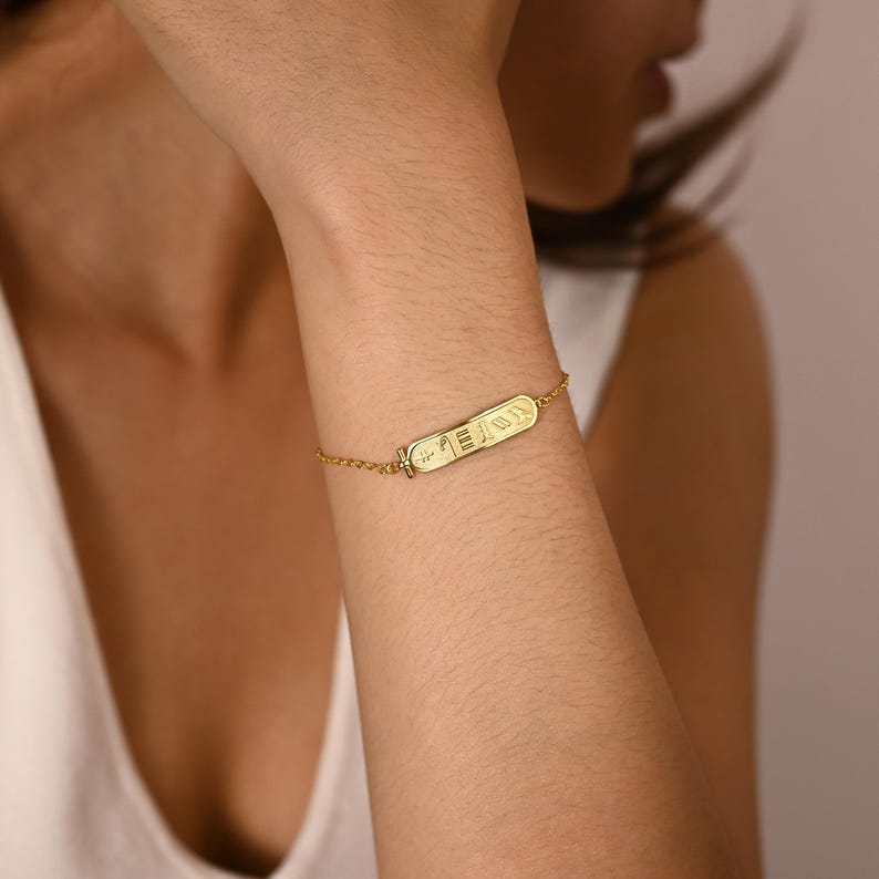 K&ouml;nnte beinhalten: Ein goldfarbenes Armband mit einem gravierten rechteckigen Anh&auml;nger mit hieroglyphischen Symbolen. Das Armband ist an einer zarten Kette befestigt und wird am Handgelenk getragen. Der Schmuck wird vor einem neutralen Hintergrund pr&auml;sentiert.