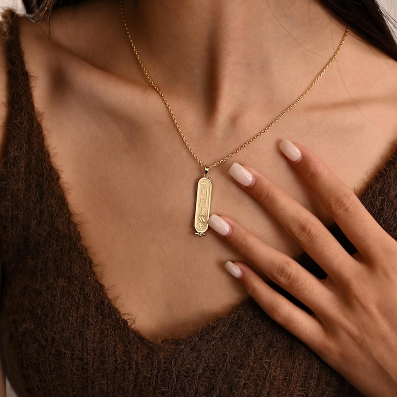 K&ouml;nnte beinhalten: Eine goldfarbene Halskette mit Anh&auml;nger, die einen l&auml;nglichen, rechteckigen Anh&auml;nger mit eingravierten Hieroglyphen-Symbolen aufweist. Die Halskette wird von einer Person mit einem braunen Oberteil getragen. Der Anh&auml;nger h&auml;ngt an einer zarten Kette.
