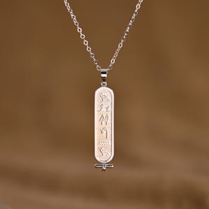 K&ouml;nnte beinhalten: Silberfarbene Halskette mit einem l&auml;nglichen, rechteckigen Anh&auml;nger mit eingravierten Hieroglyphen. Der Anh&auml;nger h&auml;ngt an einer filigranen Kette. Der Hintergrund ist in einem warmen, neutralen Farbton gehalten.