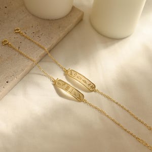 K&ouml;nnte beinhalten: Zwei goldfarbene Kettenarmb&auml;nder mit rechteckigen Anh&auml;ngern. Die Anh&auml;nger sind mit Hieroglyphen-Symbolen graviert. Die Armb&auml;nder werden auf einer wei&szlig;en Oberfl&auml;che mit neutralem Hintergrund pr&auml;sentiert, was auf ein Schmuckprodukt hindeutet.