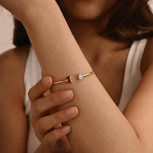 May include: Gold cuff bracelet with a rectangular, deep red gemstone and a pear-shaped, clear gemstone. The open design allows for easy wear. A simple yet elegant piece of jewellery.