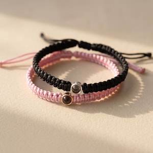 Peut inclure: Deux bracelets d'amiti&eacute; faits &agrave; la main. L'un est noir et tress&eacute;, l'autre rose et tiss&eacute;. Chaque bracelet comporte un petit charme rond. Les bracelets sont sur un fond neutre.