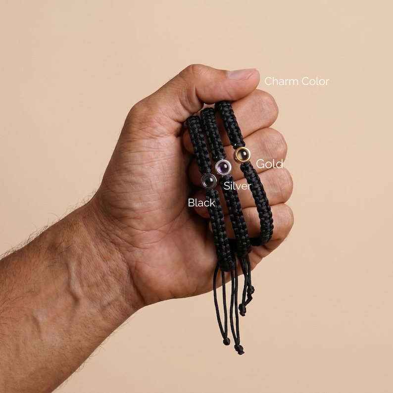 Peut inclure: Bracelets noirs tress&eacute;s avec des liens r&eacute;glables. Chaque bracelet pr&eacute;sente un charm en or, argent ou noir. Les charms sont circulaires et ont une pierre pr&eacute;cieuse au centre. Les bracelets sont tenus dans une main.