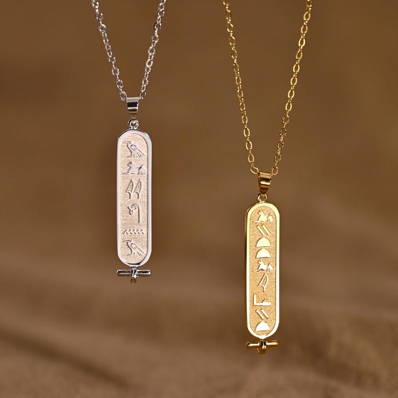 K&ouml;nnte beinhalten: Zwei Anh&auml;ngerhalsketten, eine silberne und eine goldene, jeweils mit einem l&auml;nglichen rechteckigen Anh&auml;nger. Die Anh&auml;nger weisen eingravierte Hieroglyphen-Symbole auf. Die silberne Halskette hat eine zarte Kette, w&auml;hrend die goldene Halskette eine Gliederkette hat.