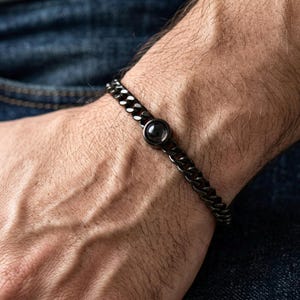 Peut inclure: Un bracelet chaîne gris foncé avec une pierre noire circulaire au centre. Le bracelet est composé de maillons et est porté au poignet. L'arrière-plan est une vue floue de denim.