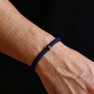 Peut inclure: Un bracelet en macram&eacute; bleu marine avec un fermoir &agrave; perle noire. Le bracelet est port&eacute; sur un poignet sur fond sombre. Le macram&eacute; est tiss&eacute; serr&eacute; et la perle est centr&eacute;e sur le bracelet.